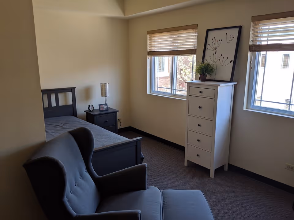 A small bedroom with a single bed, a nightstand with a lamp and a clock, a gray armchair, and a white chest of drawers with a plant and framed artwork on top. Two windows with blinds allow natural light into the room.