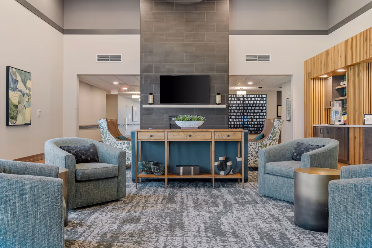 A modern and cozy common area with four light blue upholstered armchairs arranged around a wooden console table. The console table has decorative items and a plant on top. Behind the table is a dark tiled wall with a mounted flat-screen TV and two candle holders. The room features a patterned carpet, neutral walls, and a wooden accent area on the right side with shelves and lighting.