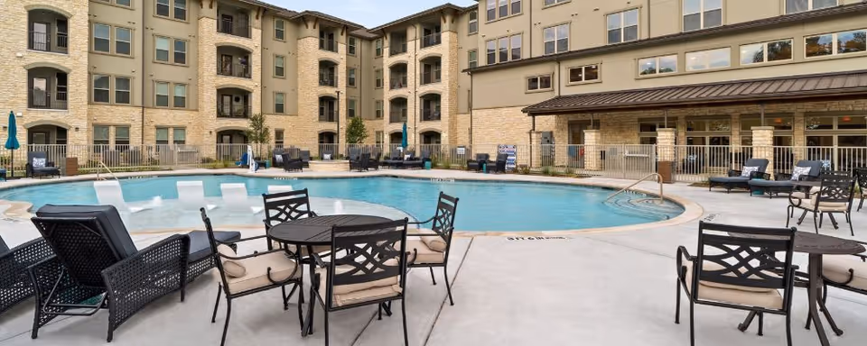Outdoor pool and patio area with tables, lounge chairs, and a multi-story apartment building in the background.