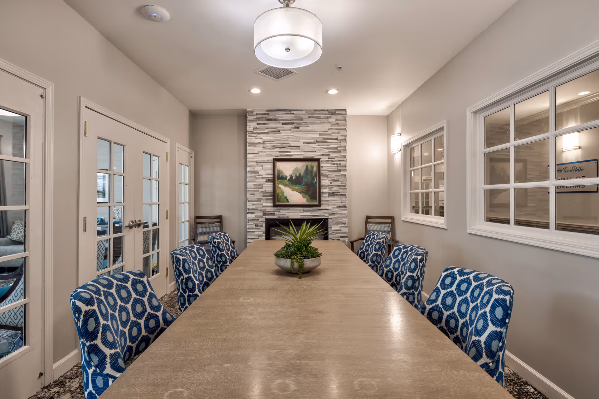 Long dining table with blue patterned chairs facing a stone fireplace and framed artwork in a modern interior room.