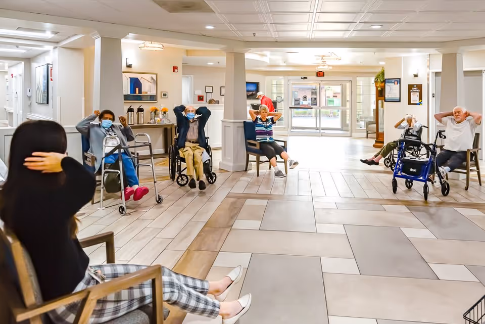 A group of elderly individuals seated in chairs and wheelchairs in a spacious, well-lit common area of a senior living facility, participating in a seated exercise session led by a woman in the foreground. The room features tiled flooring, white walls, and large windows near the entrance letting in natural light.
