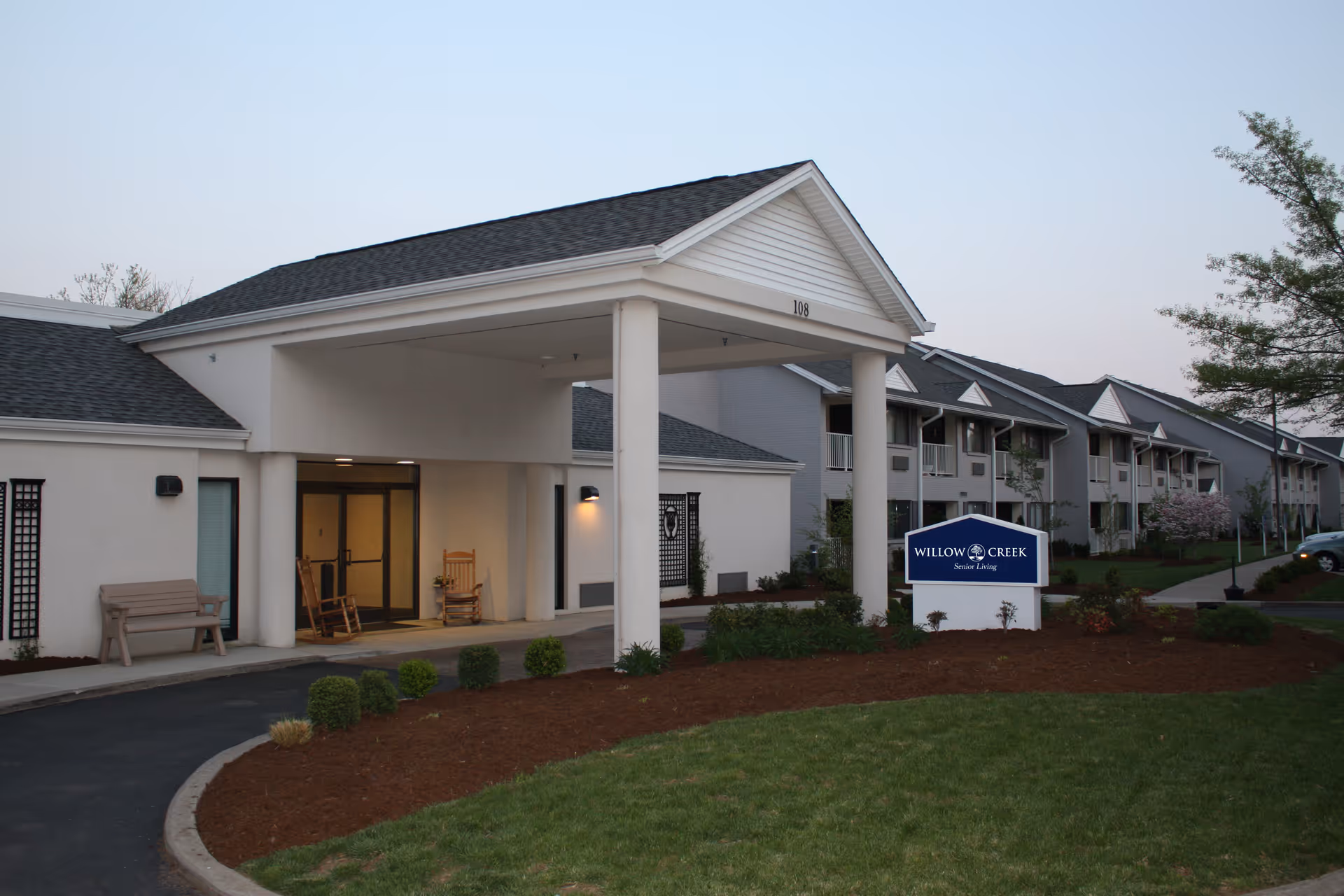Exterior view of Willow Creek Senior Living facility showing the entrance with a covered drop-off area supported by columns, a bench and rocking chairs near the entrance, landscaped garden beds, and a sign with the facility name in front of a multi-story building.