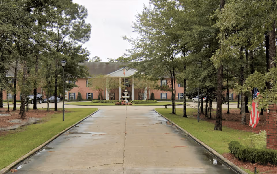 A wide concrete driveway lined with trees and lamp posts leads to a large brick building with a white columned entrance and a fountain in front. The area is surrounded by greenery and appears to be part of a senior living facility.