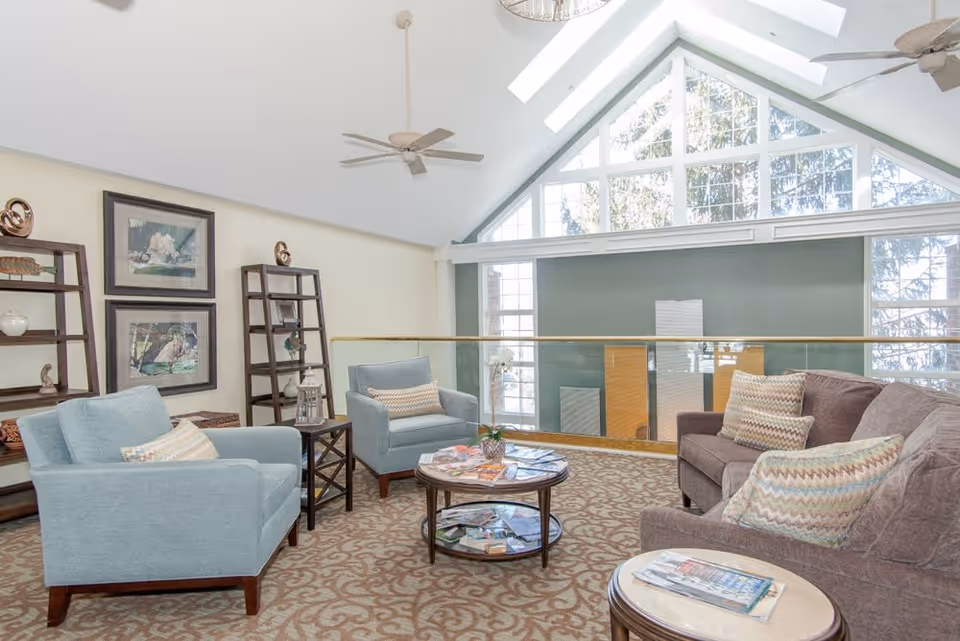 A bright and spacious living room area with large triangular windows letting in natural light. The room features two light blue armchairs and a brown sofa, all with patterned throw pillows. There are two round coffee tables with magazines on them, and two wooden shelving units with decorative items. The ceiling has two ceiling fans and skylights.