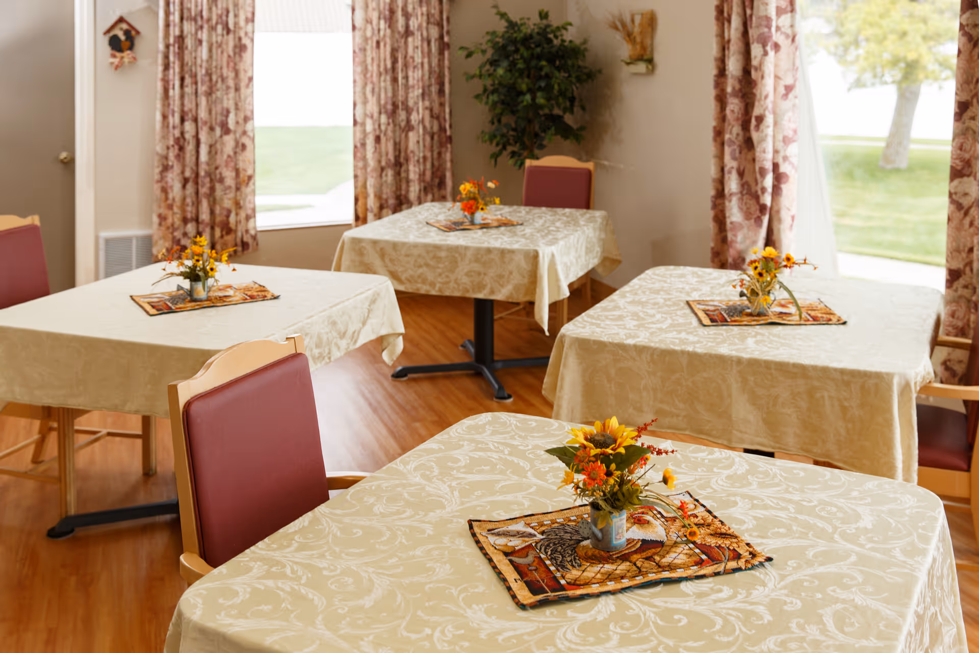 Dining room with multiple tables draped in light patterned tablecloths, small floral centerpieces, upholstered chairs, and windows with floral curtains.