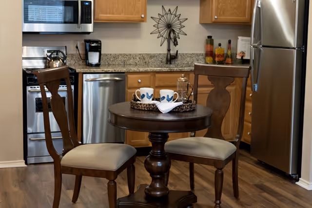 A small kitchen area with wooden cabinets, granite countertops, and stainless steel appliances including a microwave, dishwasher, stove, and refrigerator. In front of the kitchen is a round wooden table with two wooden chairs, each with cushioned seats. On the table are two coffee mugs, a French press, and a napkin on a woven tray.