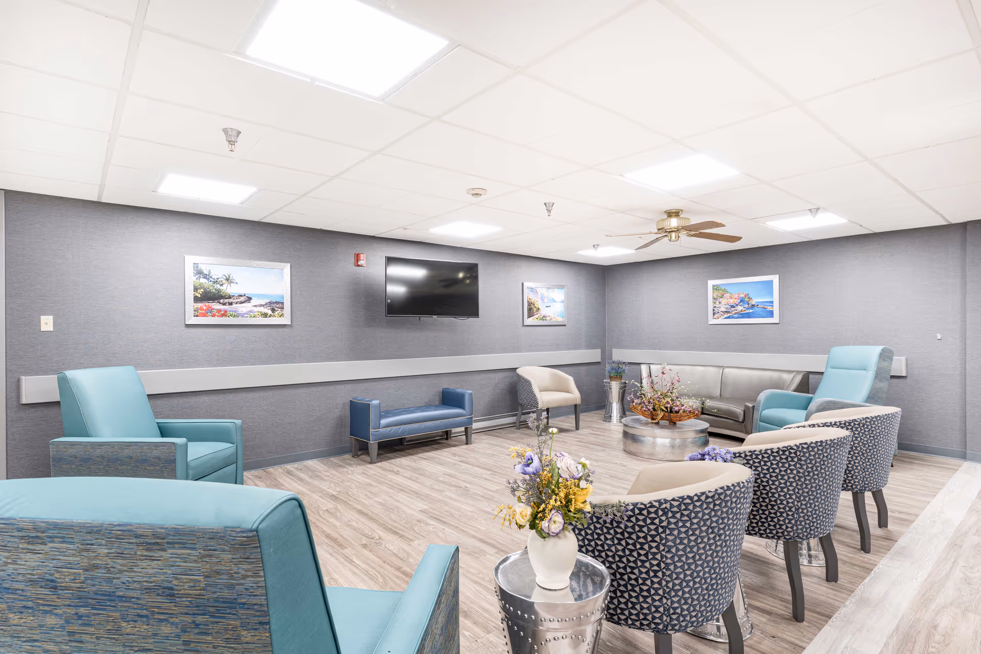 A bright and spacious common area in a senior living facility with modern furniture including teal armchairs, patterned chairs, a gray sofa, and a blue bench. The room has light wood flooring, gray textured walls adorned with framed landscape pictures, a ceiling fan, and a wall-mounted flat-screen TV. There are decorative flower arrangements on metal side tables and a round coffee table in the center.