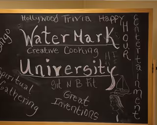 A chalkboard with various words and phrases written in white chalk, including 'WaterMark University' prominently in the center. Other words around the board include 'Hollywood Trivia', 'Happy Hour', 'Creative Cooking', 'Entertainment', 'Spiritual Gathering', 'Sit N B Fit', and 'Great Inventions'. There is also a small drawing of a sailboat and a face on the right side.