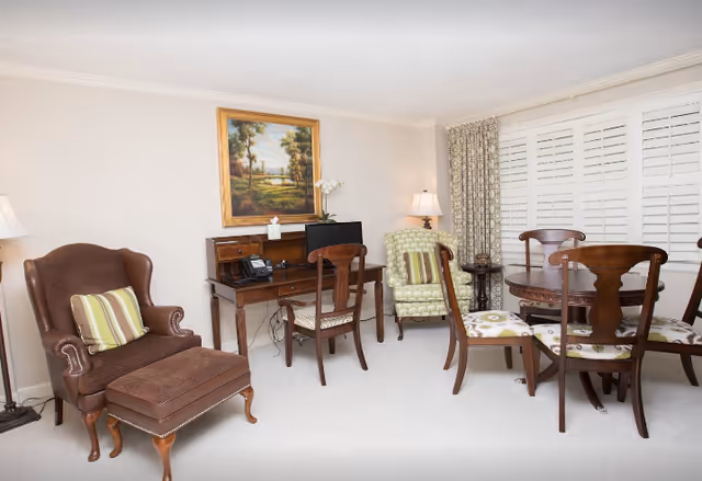 Bright living room with upholstered armchairs, a small wooden desk, and a round dining table with chairs in front of shuttered windows.