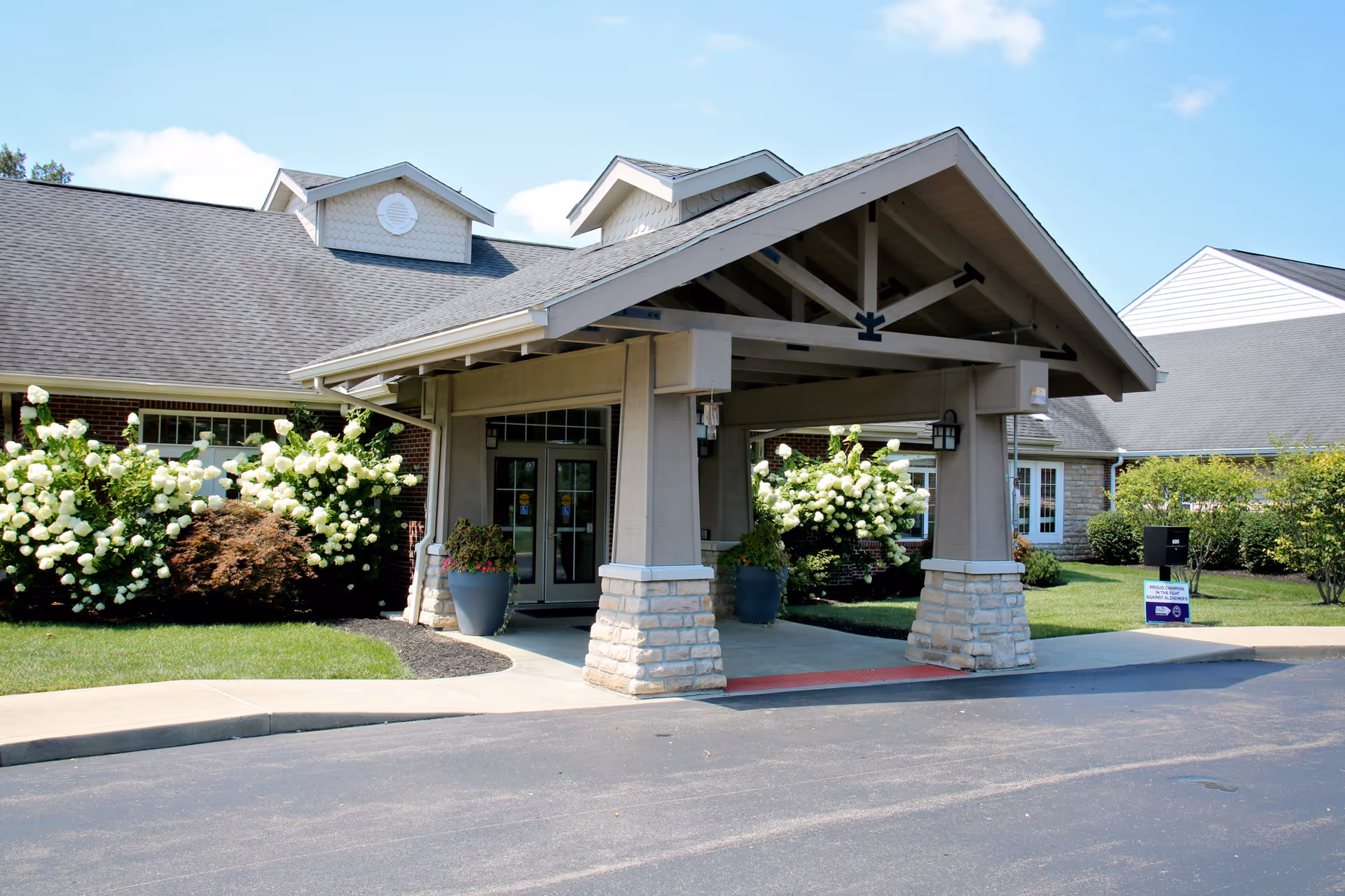 Entrance of a senior living facility named Legacy Village with a covered drop-off area supported by stone and wood pillars, surrounded by green grass, white flowering bushes, and a clear blue sky.