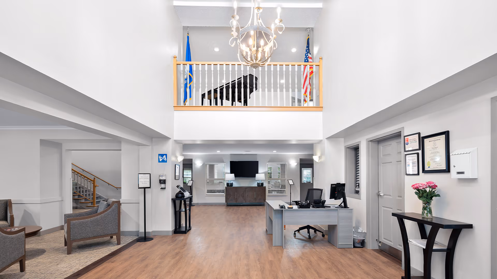 Bright and spacious reception area of a senior living facility with a wooden floor, a desk with computer and office chair on the right, seating area with chairs on the left, a staircase leading upstairs, and a balcony above with a grand piano and two flags. A chandelier hangs from the ceiling.