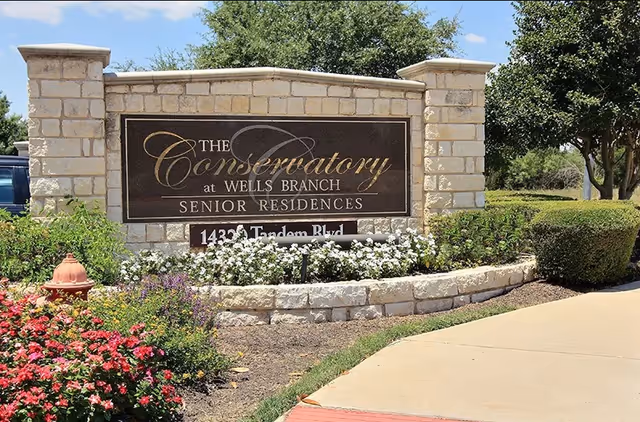 Stone sign for The Conservatory at Wells Branch Senior Residences surrounded by flowers and greenery, with a sidewalk and trees in the background under a blue sky.