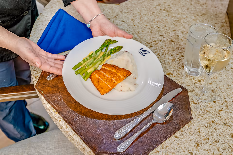 A person presenting a plate of food on a table with a brown placemat. The plate contains cooked salmon, mashed potatoes with gravy, and steamed asparagus. There is a glass of water and a glass of white wine on the table, along with a knife and spoon.