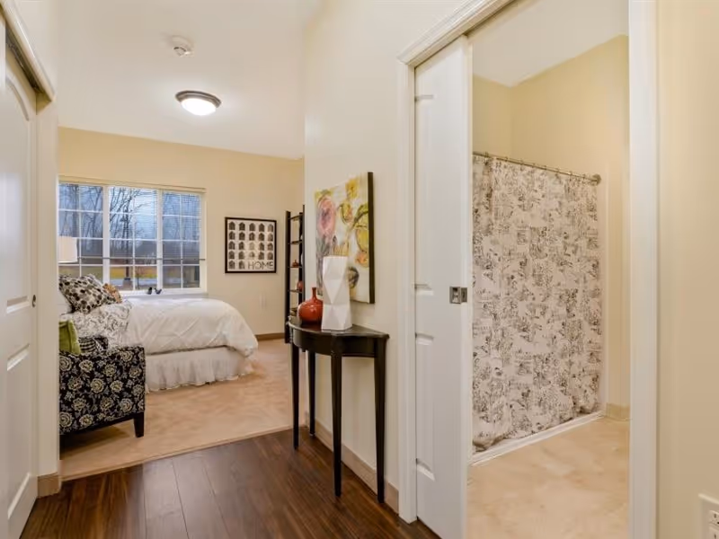 View of a senior living facility room showing a hallway with dark wood flooring leading to a bedroom on the left with a bed, patterned armchair, window, and wall art. On the right, there is a bathroom with a shower curtain featuring a black and white pattern.