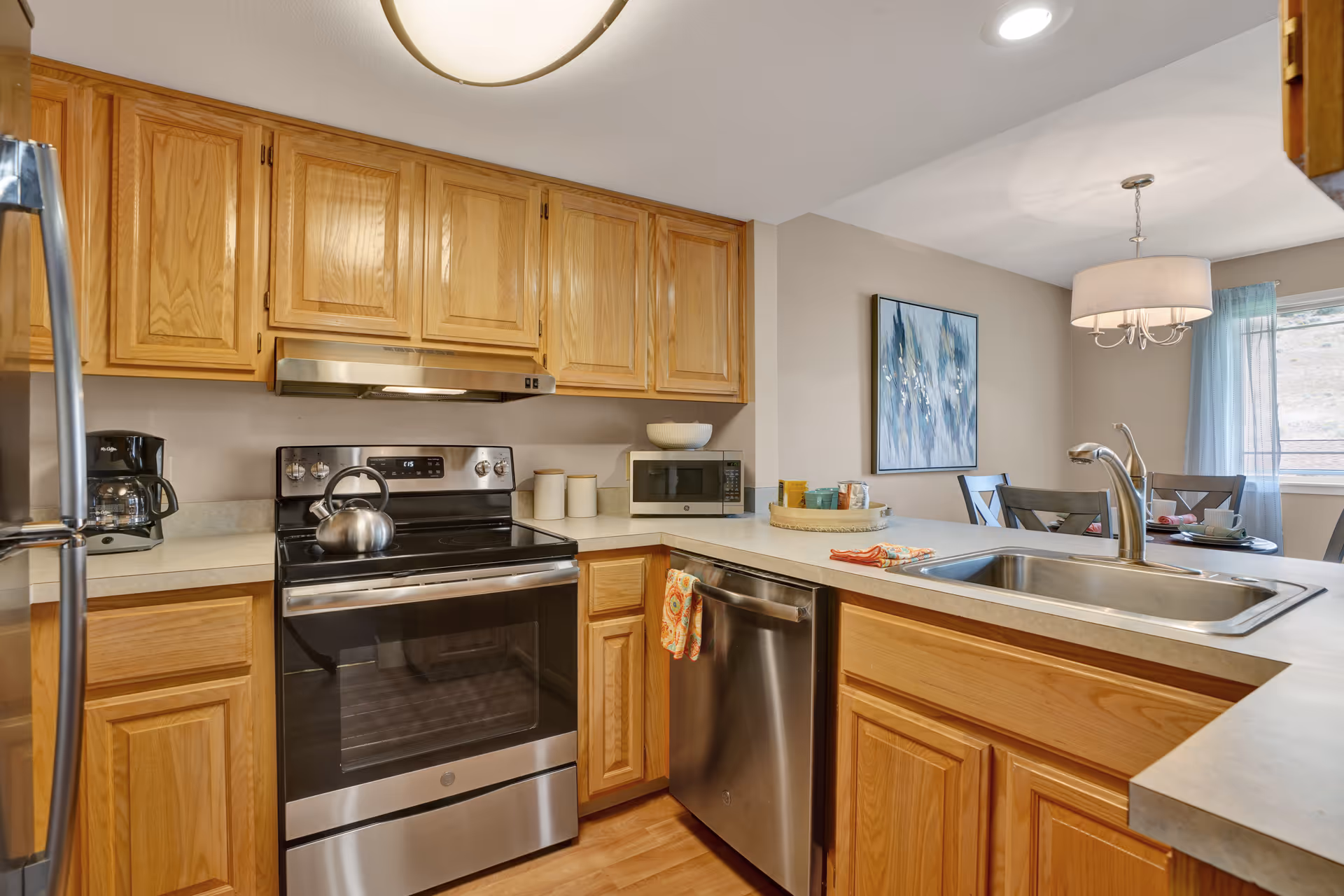 A modern kitchen with wooden cabinets, a stainless steel oven with a kettle on top, a dishwasher, a microwave, and a coffee maker. The kitchen counter has a sink and some colorful towels hanging. In the background, there is a dining area with a table, chairs, a hanging light fixture, a window with sheer curtains, and a blue abstract painting on the wall.