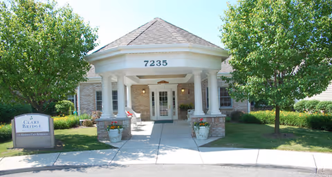 Front entrance of a brick senior living building with a circular covered portico marked 7235, flanked by columns, planters, and trees.