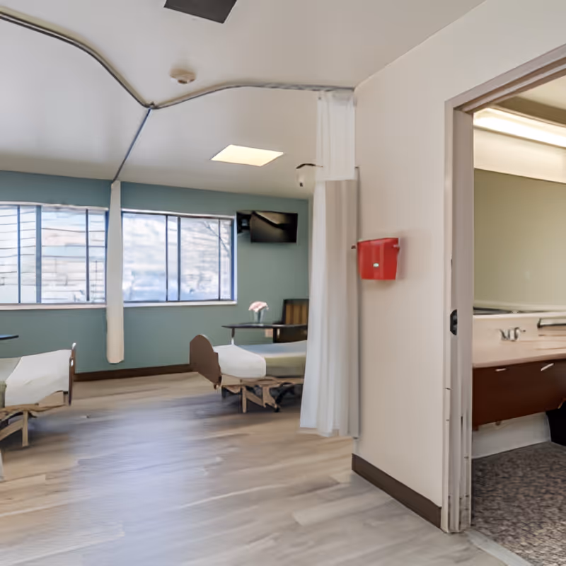 Shared resident room with two adjustable beds, privacy curtains, a wall-mounted TV, and a sink/vanity area visible through a doorway.