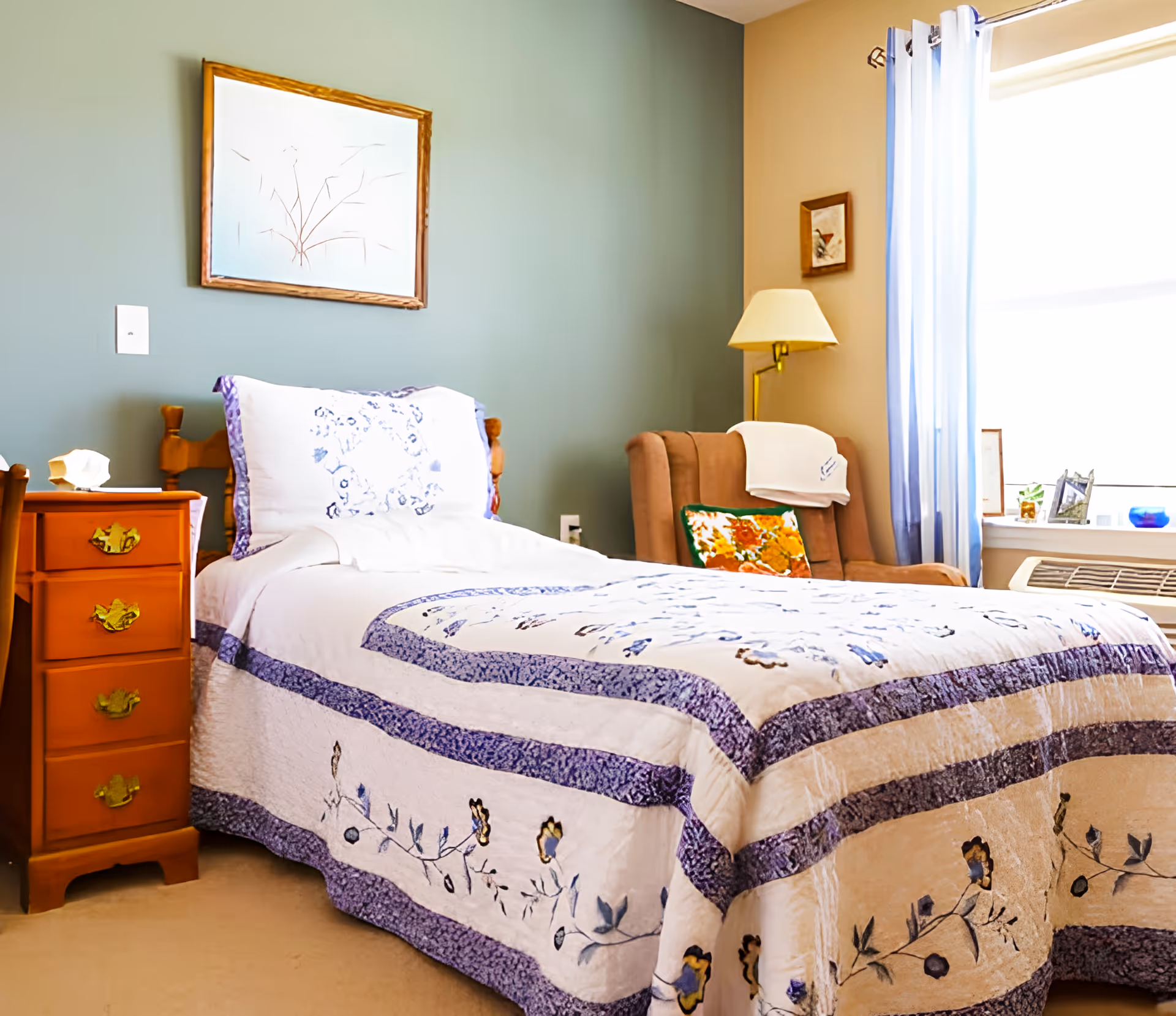 A cozy bedroom with a single bed covered in a white and purple floral quilt. Next to the bed is a wooden nightstand with drawers and a seashell decoration. A cushioned armchair with a colorful pillow and a white cloth draped over the back is positioned near a window with sheer white curtains. A floor lamp and framed artwork hang on the walls painted in muted green and beige tones.