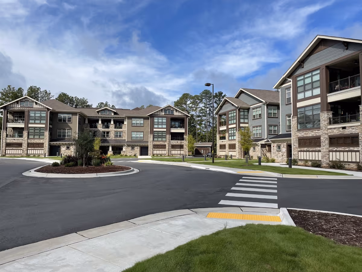 Exterior view of three-story apartment-style senior living buildings with balconies, a landscaped roundabout, and a pedestrian crosswalk.