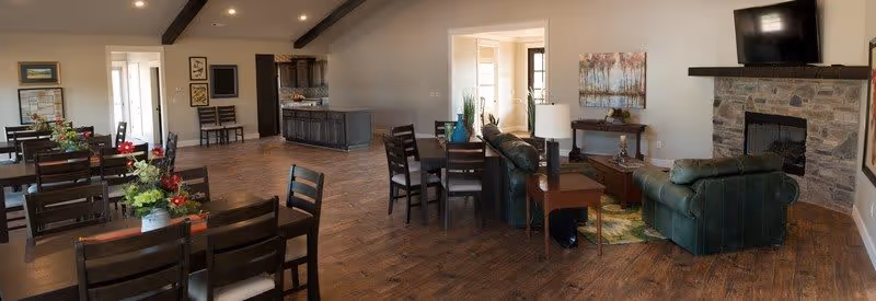 Spacious interior room with wooden flooring featuring multiple dining tables with chairs and floral centerpieces on the left side, and a cozy seating area with green leather sofas, a wooden coffee table, a side table with a lamp, a stone fireplace with a mounted TV above it, and wall art on the right side.