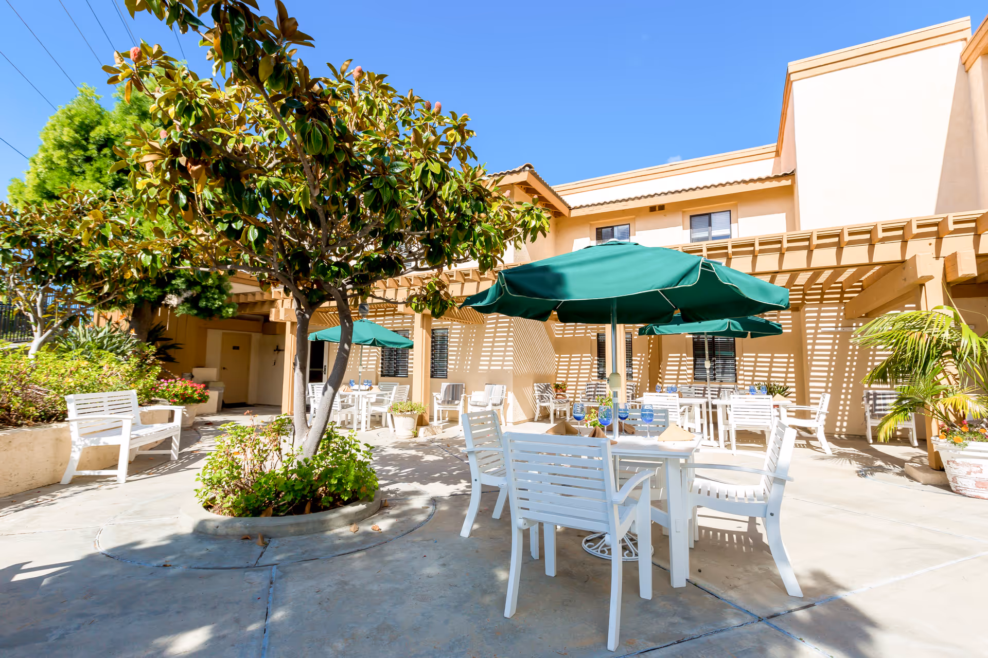 Outdoor patio area at Brookdale Senior Living with white tables and chairs, green umbrellas providing shade, surrounded by trees and plants, under a clear blue sky.