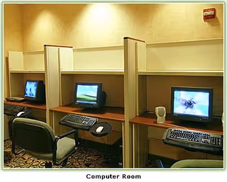 A computer room with three desktop computers placed in individual cubicles. Each cubicle has a chair, keyboard, and mouse. The room has beige walls and carpeted flooring with a patterned design.