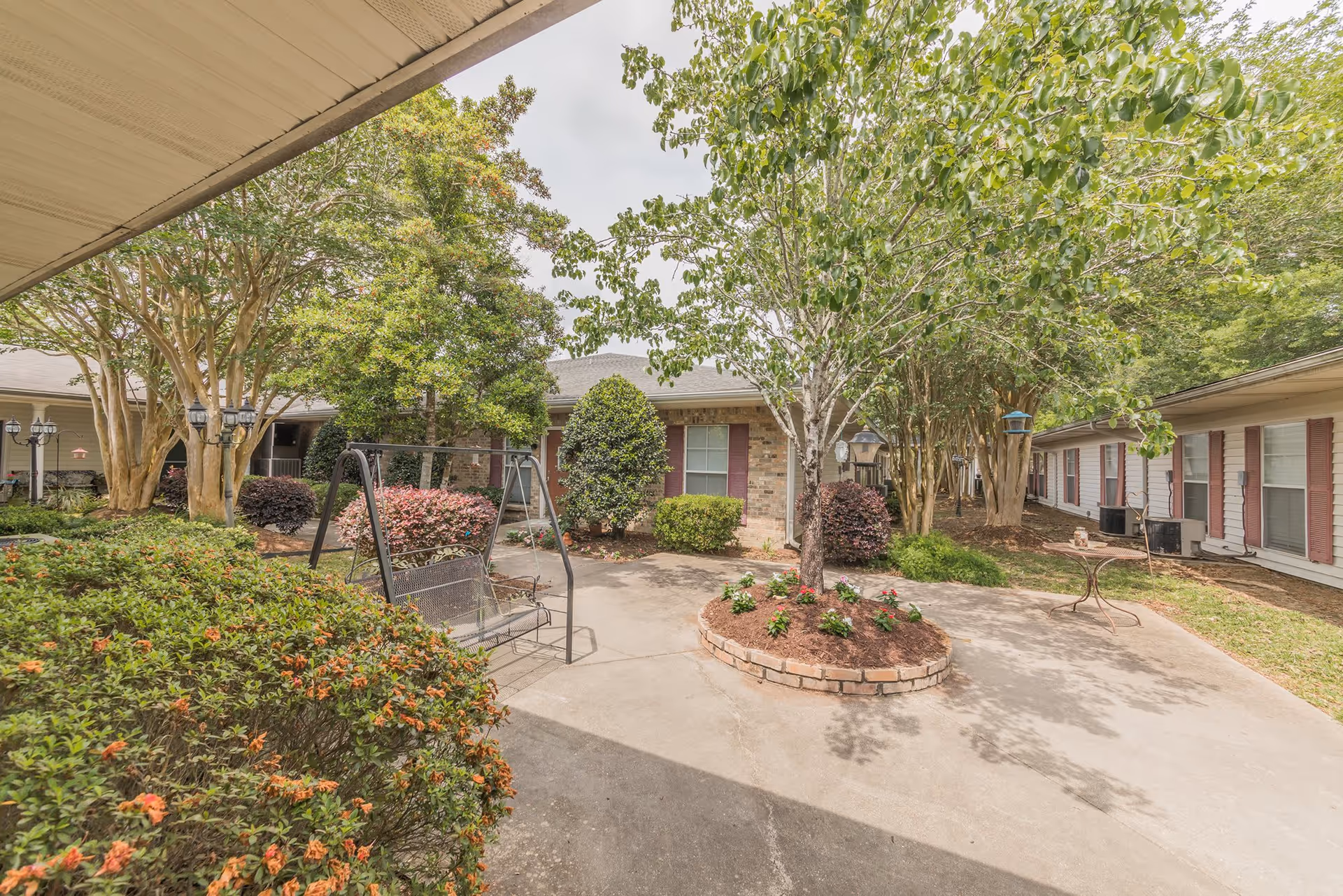 Outdoor courtyard area at Rosewood Assisted Living featuring a concrete patio with a metal swing bench, surrounded by various trees, bushes, and flower beds. The courtyard is enclosed by single-story buildings with windows and red shutters.