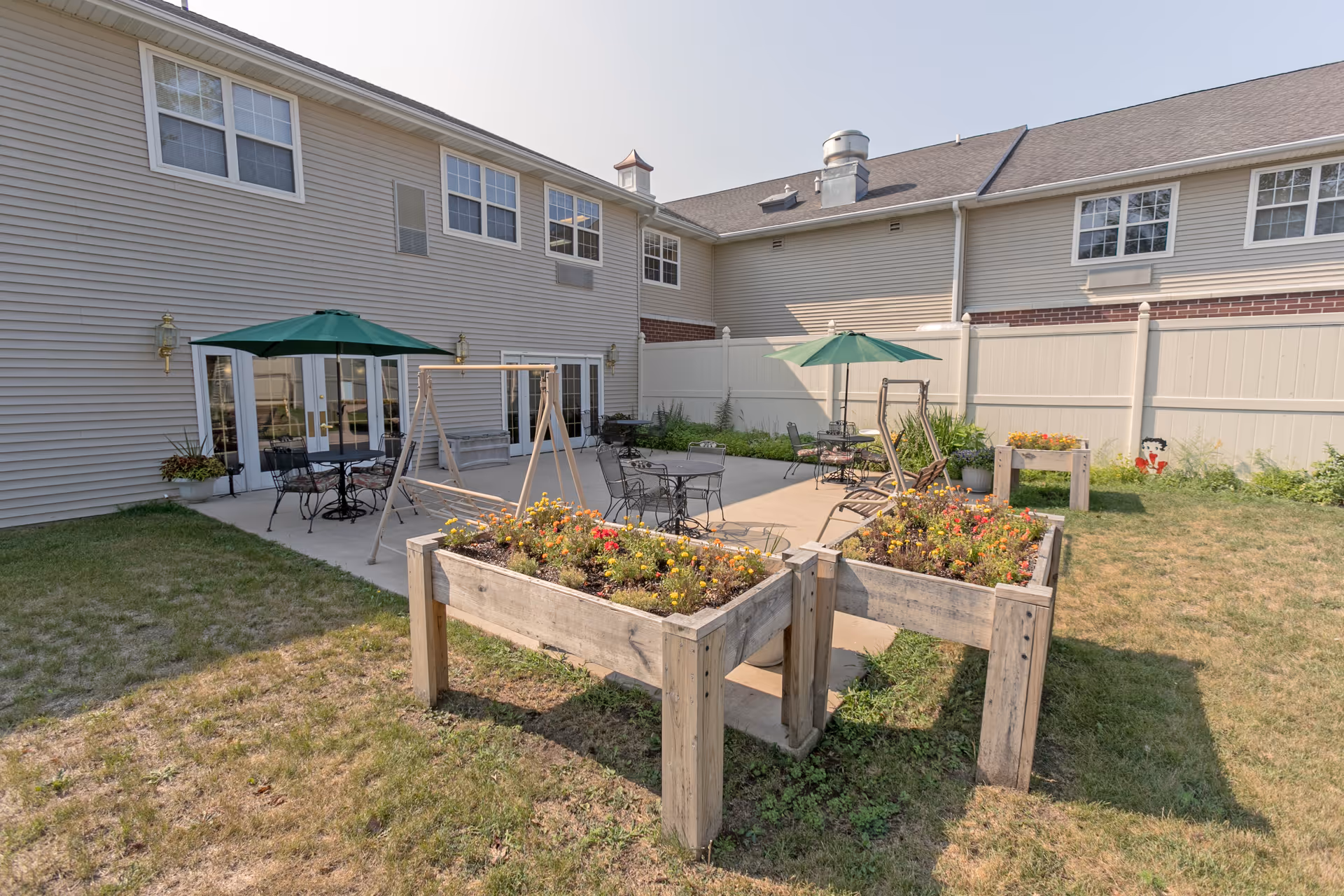 Outdoor patio area at Heritage Woods of Manteno featuring raised garden beds with flowers, metal tables and chairs with green umbrellas, a wooden swing, and a beige privacy fence surrounding the space. The building exterior is light-colored with multiple windows and a door leading to the patio.