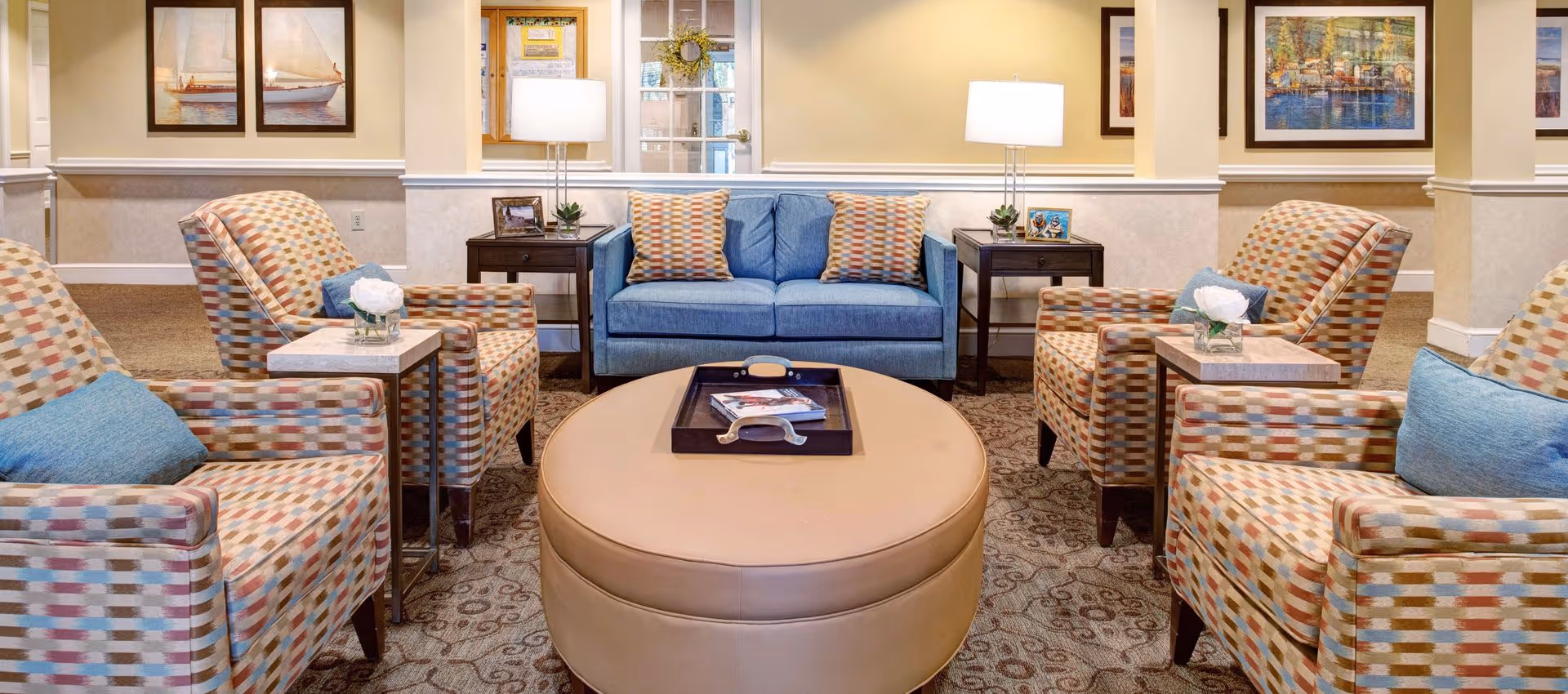 A cozy living room area with a blue sofa adorned with patterned cushions, four patterned armchairs with blue pillows, two side tables with lamps and small plants, a round beige ottoman with a tray and magazines on top, and framed artwork on the walls.
