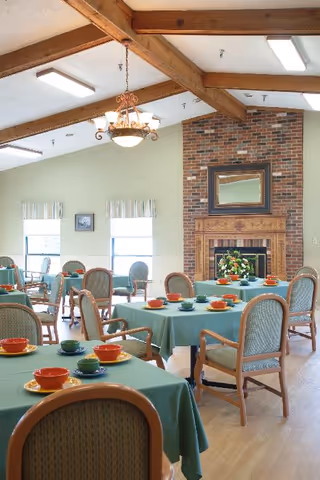 A dining room with multiple tables covered in green tablecloths, each set with colorful bowls and plates. The room features wooden chairs with cushioned seats, a brick fireplace with a mirror above it, large windows with striped valances, exposed wooden ceiling beams, and a chandelier hanging from the ceiling.
