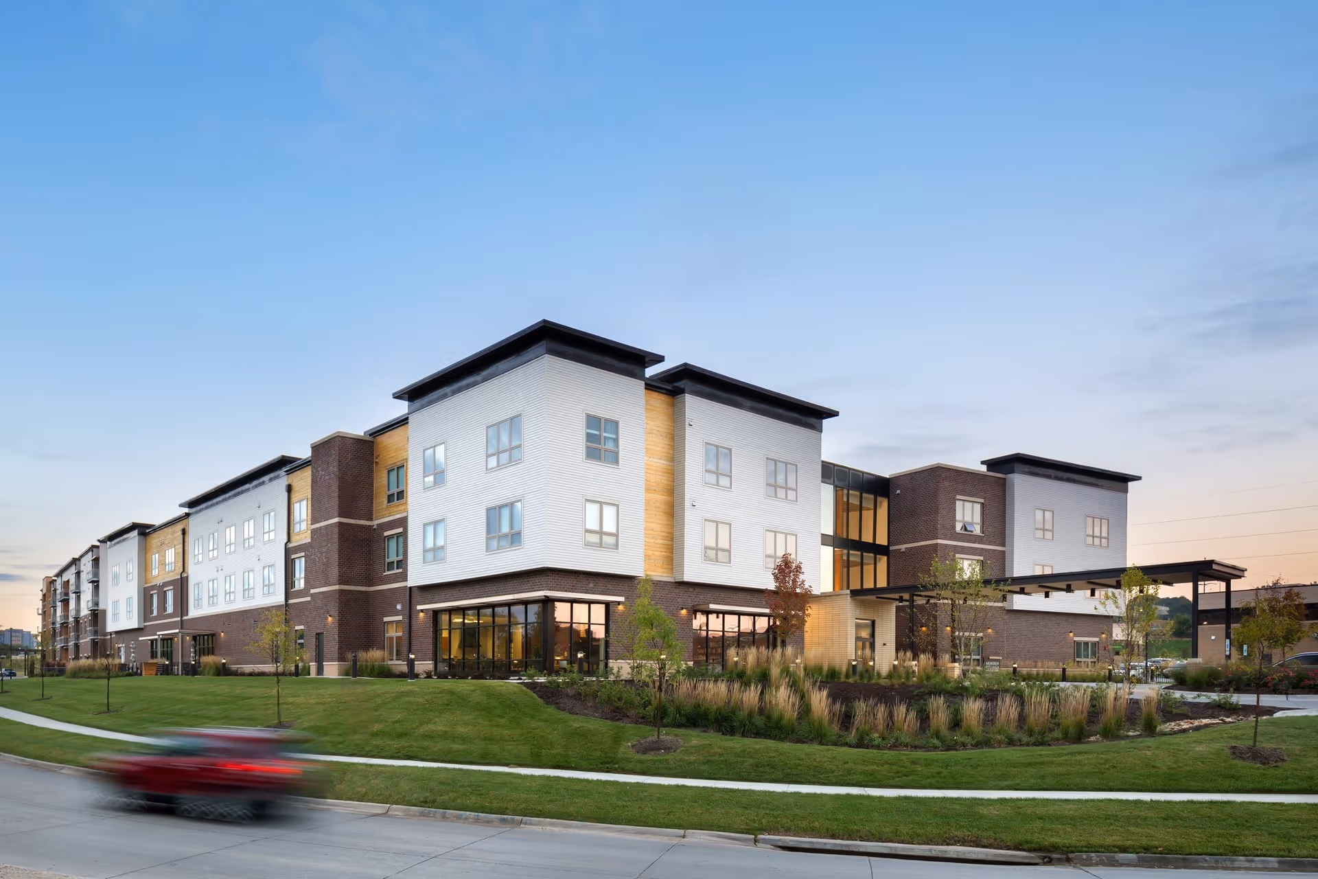 Exterior view of a modern multi-story senior living facility building with large windows, landscaped greenery, and a clear sky at dusk. A red car is blurred in motion on the road in front of the building.