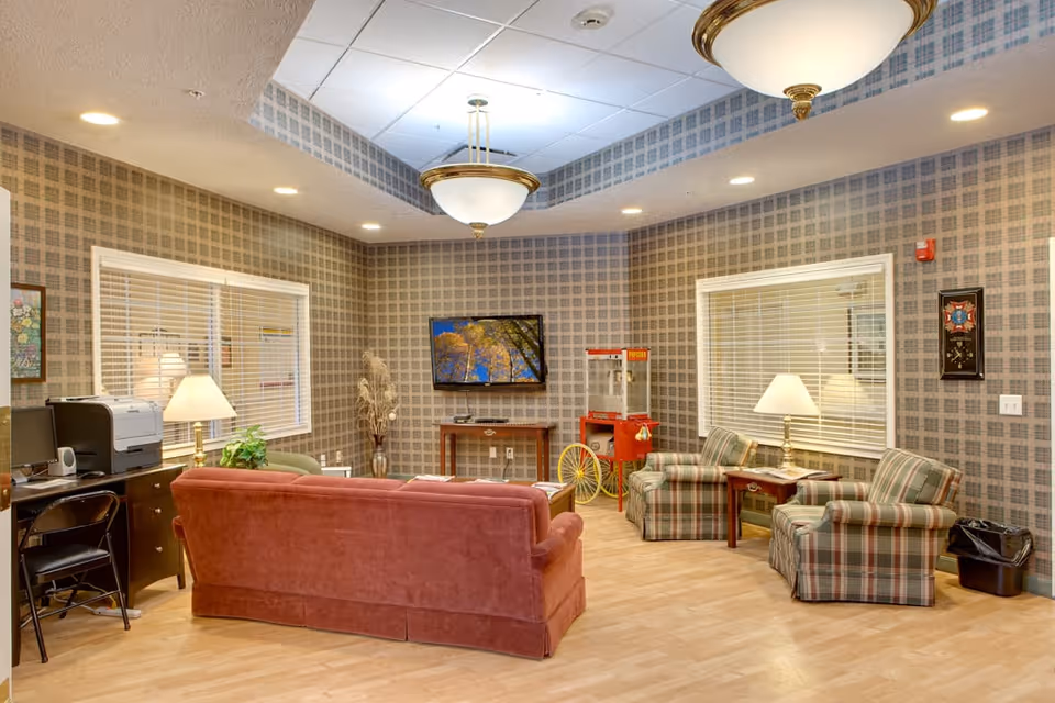 A cozy living room area with a red sofa, two plaid armchairs, a wooden coffee table, and a TV mounted on the wall. The room has patterned wallpaper, two windows with blinds, ceiling lights, and a popcorn machine in the corner. There is also a desk with a printer and a chair on the left side.