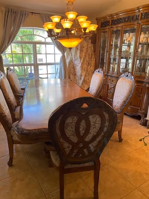 A formal dining room with a long wooden dining table surrounded by eight upholstered chairs. A large chandelier with multiple lights hangs above the table. There is a large window with curtains on the left side, and a wooden china cabinet with glass doors on the right side displaying glassware.