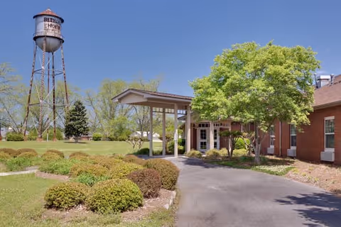 Exterior view of The Oaks - Bethany Assisted Living facility showing a driveway leading to a covered entrance with a tree and bushes on the right side. A tall water tower labeled 'Bethany Home' is visible on the left against a clear blue sky.
