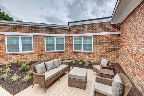 Outdoor patio area with wicker furniture including a sofa, two armchairs, a small table, and an ottoman. The patio is surrounded by brick walls with several windows and has a wooden deck floor. There are small plants and mulch beds along the edges of the patio.