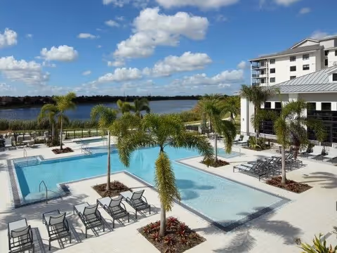 Outdoor swimming pool area with several palm trees and lounge chairs arranged around the pool. In the background, there is a large body of water and a multi-story building under a partly cloudy blue sky.