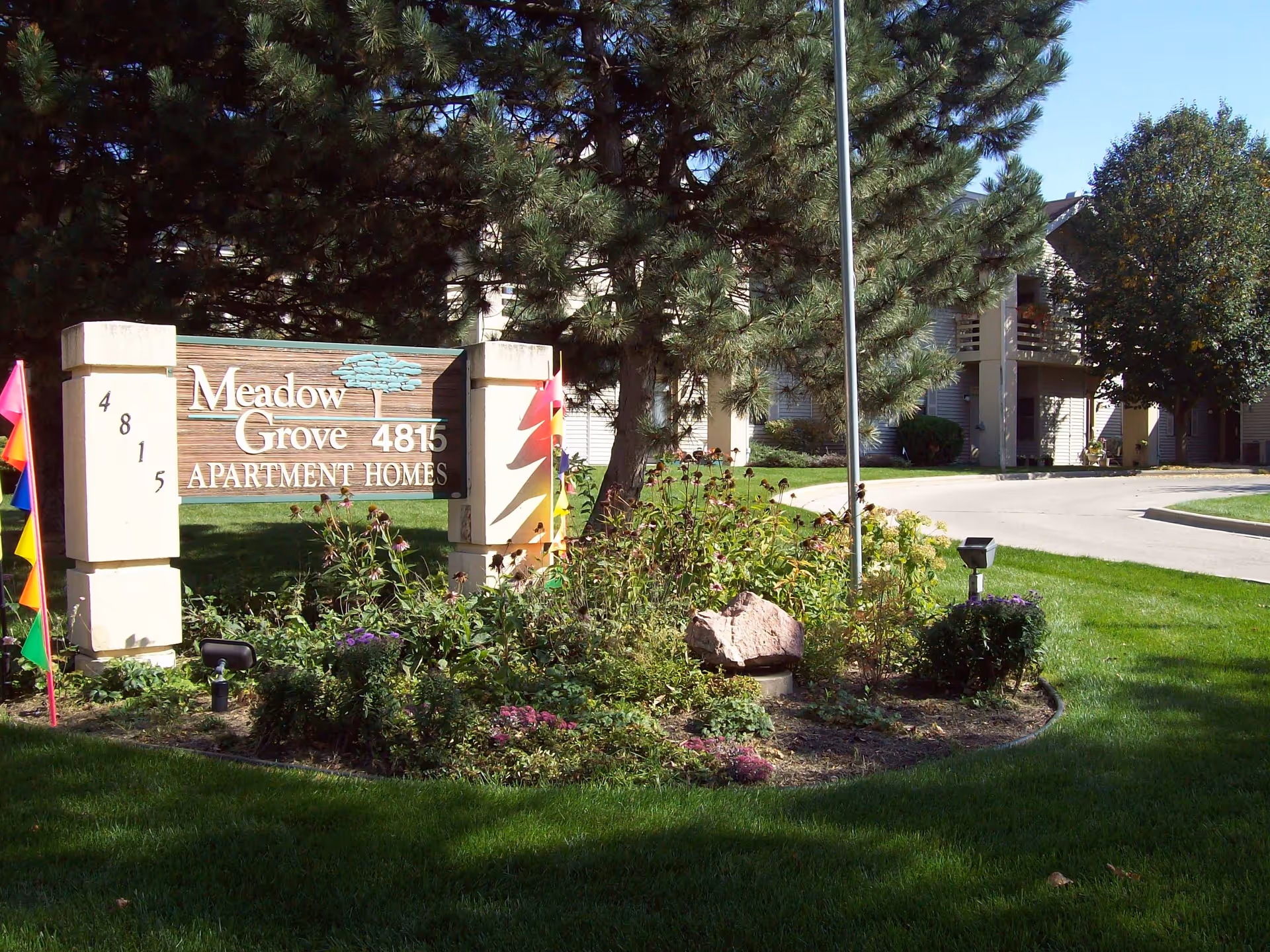 Outdoor view of the entrance sign for Meadow Grove Apartment Homes at 4815, surrounded by green grass, plants, flowers, and trees with apartment buildings visible in the background.
