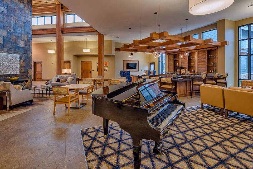 Spacious and well-lit common area featuring a black grand piano on a patterned rug, multiple seating arrangements with chairs and sofas, a stone accent wall with a decorative piece, wooden beams, and a bar area with high stools and pendant lighting.