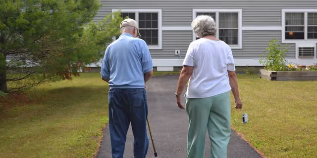 An elderly man and woman walking on a paved path beside a building with gray siding and white-framed windows. The man is using a cane and wearing a light blue shirt and dark pants, while the woman is wearing a white shirt and light green pants, holding a small object in her right hand. There is grass and a small tree on the left side of the path.