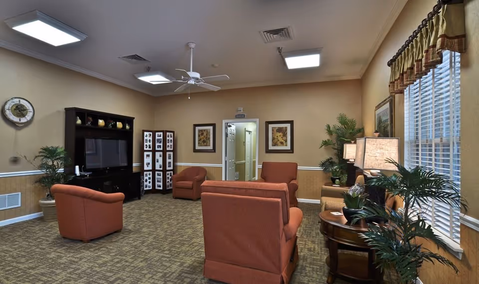 A cozy living room in a senior living facility with orange upholstered chairs arranged around a TV on a black entertainment center. The room has beige walls, carpeted floor, ceiling fan, framed artwork, potted plants, and a window with blinds and a valance.