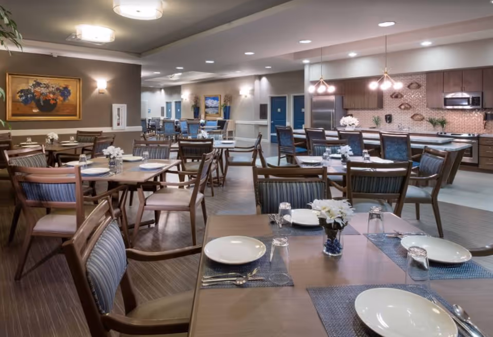 A spacious dining room in a senior living facility with multiple wooden tables and chairs arranged neatly. Each table is set with white plates, glasses, silverware, and small floral centerpieces. The room features modern lighting fixtures, a kitchen area with stainless steel appliances, and decorative paintings on the walls.