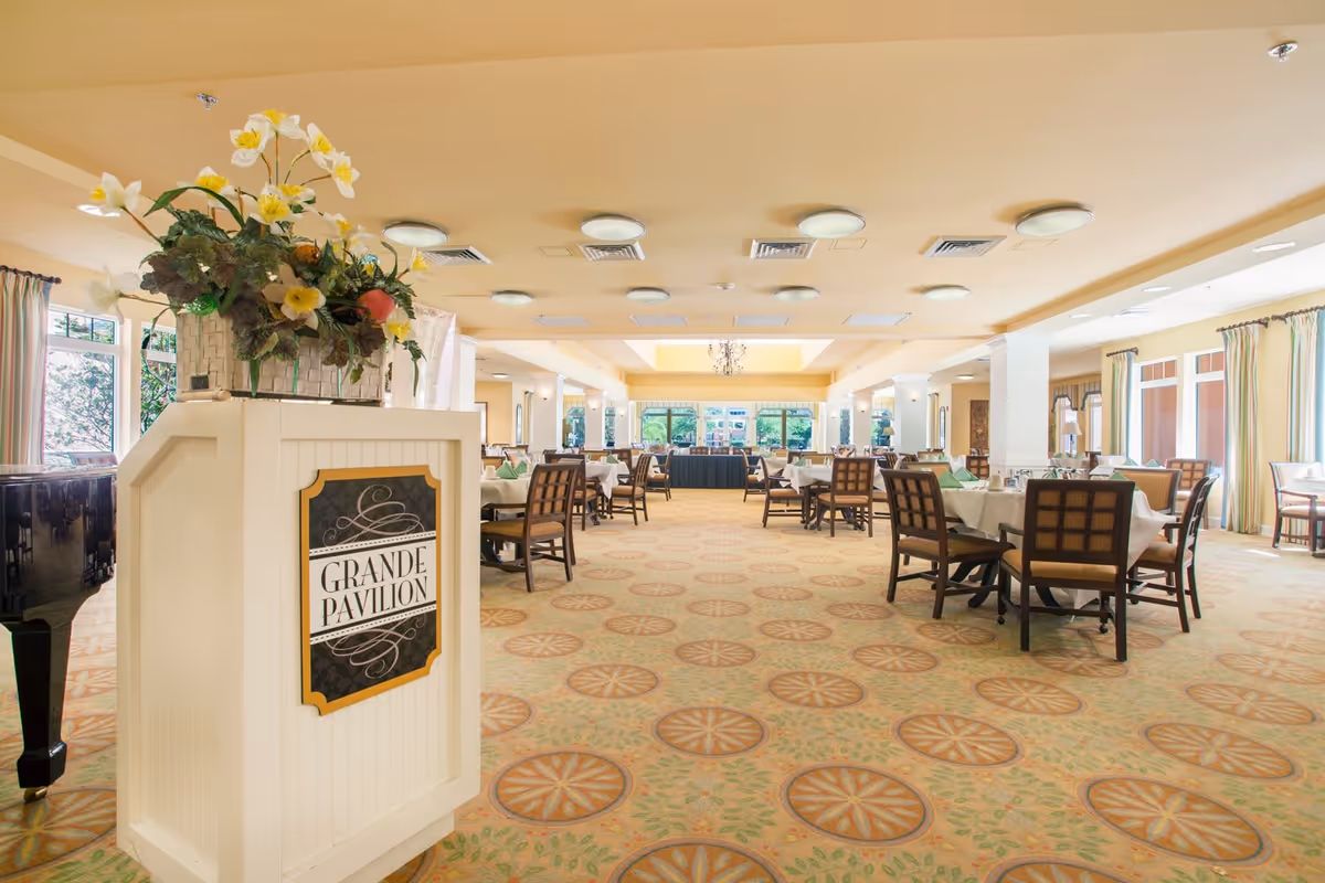 A spacious and well-lit dining room with multiple tables covered in white tablecloths and set with green napkins. The room features patterned carpet flooring, large windows with curtains allowing natural light, and a decorative podium with a flower arrangement labeled 'Grande Pavilion'.