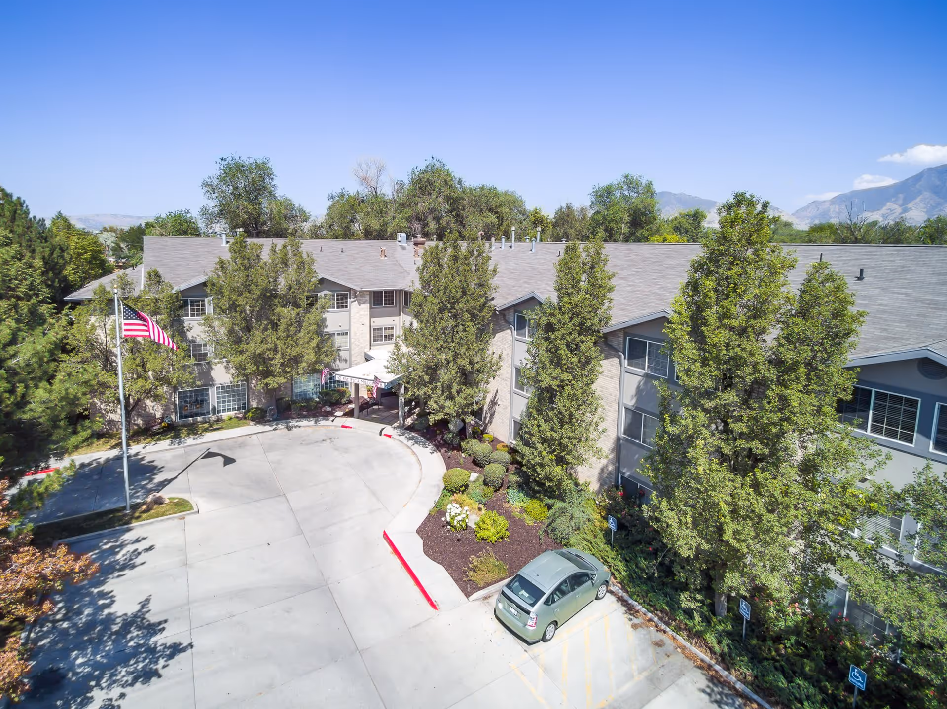 Aerial view of The Laurel at Millcreek's front entrance showing a three-story building, circular driveway, flagpole, parked car, and surrounding trees and landscaping.