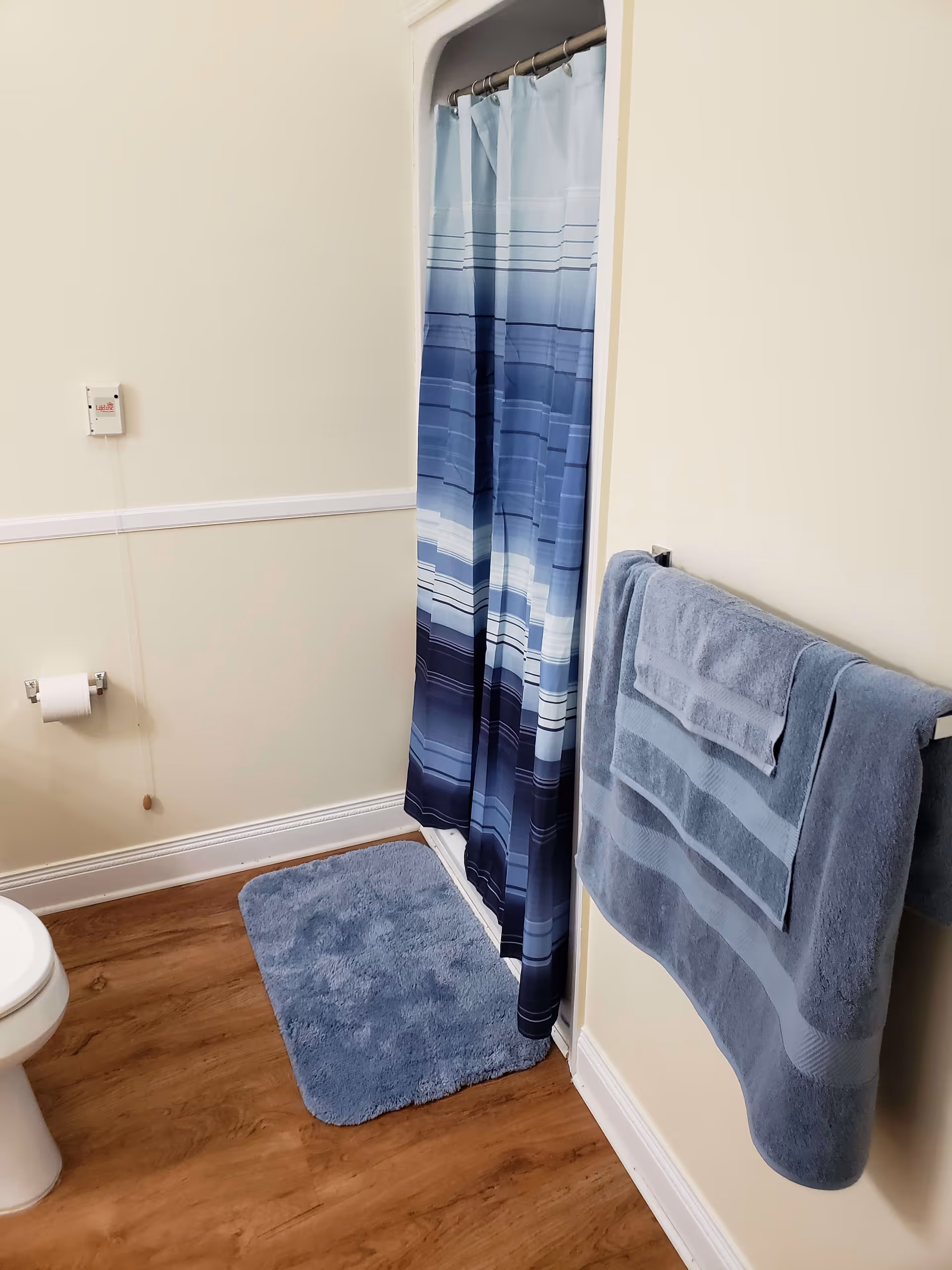 Bathroom with a white toilet, a blue and white striped shower curtain, a blue bath mat on a wooden floor, and blue towels hanging on a towel rack against a cream-colored wall.