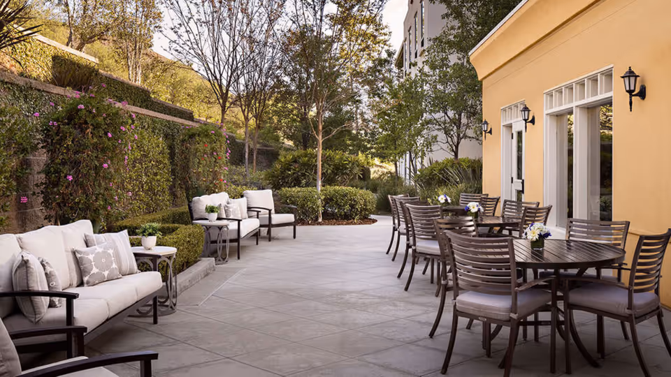 Outdoor patio area at Belmont Village Senior Living Thousand Oaks with cushioned seating on the left and multiple round tables with chairs on the right, surrounded by greenery and trees, adjacent to a yellow building with wall-mounted lanterns.