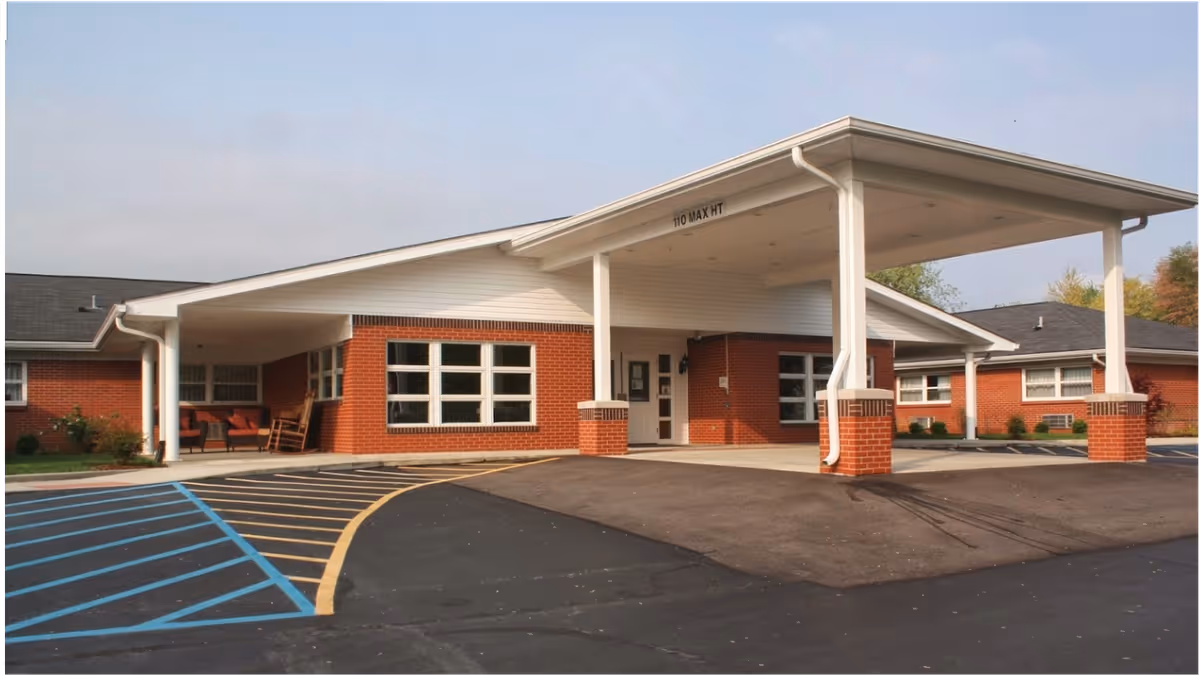 Exterior view of a single-story brick building with a covered entrance supported by white columns. The entrance area has a driveway with marked parking spaces, including a handicap parking spot. There are windows along the building and some outdoor seating visible under the covered area.