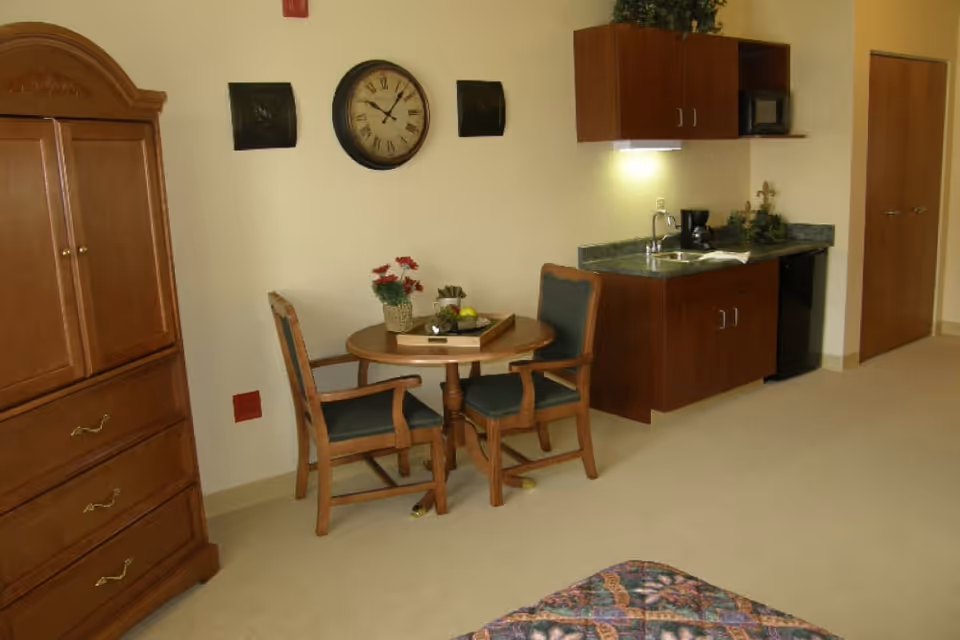 Studio-style senior living bedroom with a small round dining table and chairs, kitchenette, armoire, and a partially visible bed.