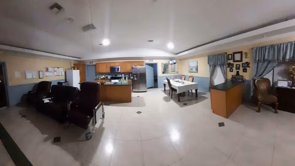 Interior view of a senior living facility common area showing a kitchen with wooden cabinets, a refrigerator, and a stove. In front of the kitchen is a dining table with chairs and a white tablecloth. To the left, there are several dark-colored recliner chairs arranged in a row. On the right side, there is a small desk area with a chair and window with blue curtains. The floor is tiled and the walls are painted in light colors with some framed pictures and bulletin boards.