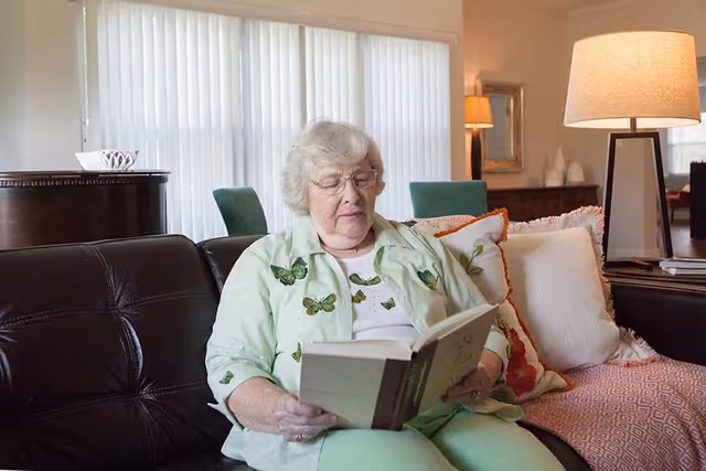 An elderly woman with gray hair and glasses is sitting on a dark leather couch in a well-lit living room, reading a book. She is wearing a light green shirt with butterfly prints and light green pants. Behind her, there are large vertical blinds covering the windows, a floor lamp, and some decorative pillows on the couch.
