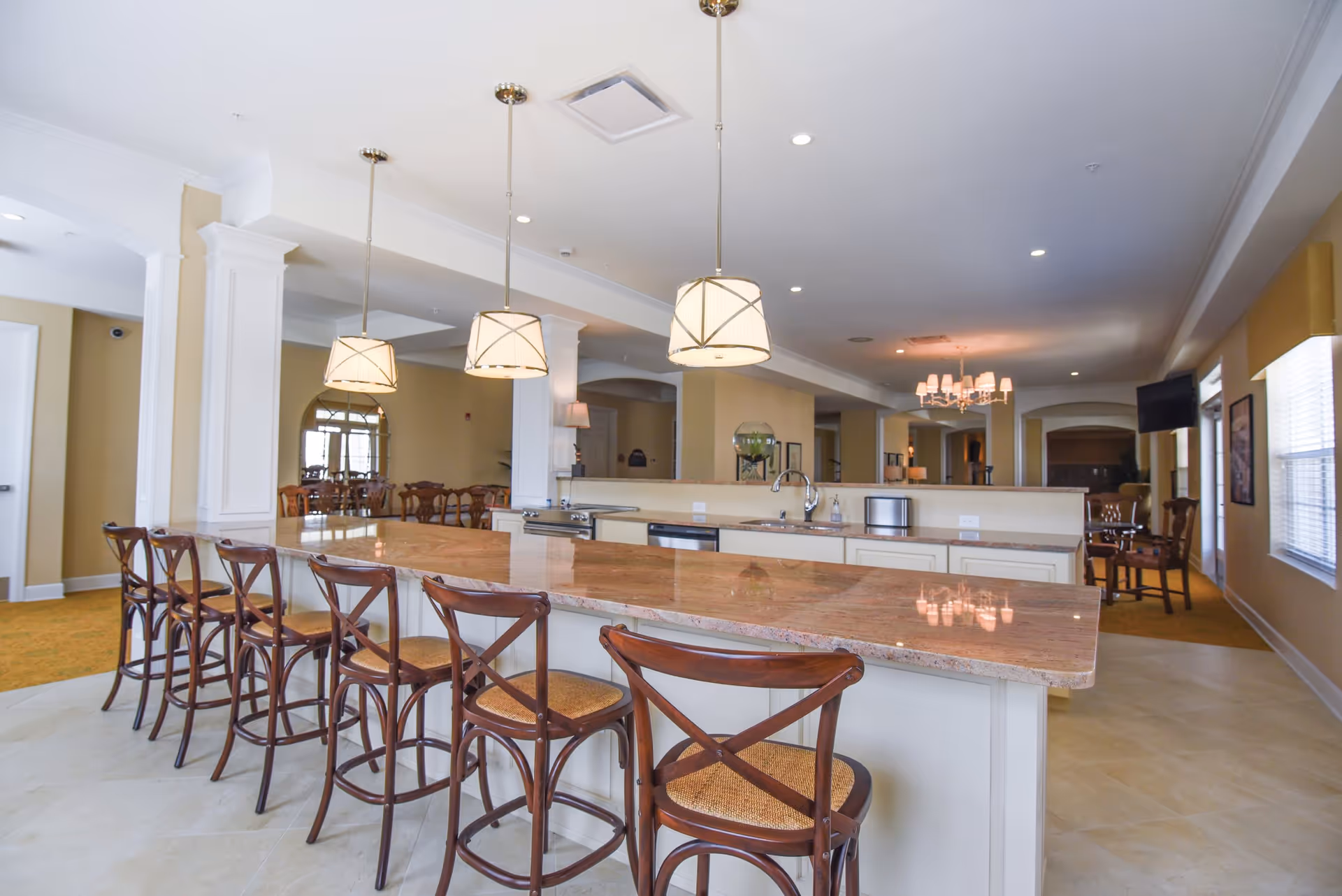 A spacious senior living facility kitchen area with a long marble countertop island surrounded by six wooden bar stools. Three pendant lights hang above the island. In the background, there are dining tables and chairs, a chandelier, and a wall-mounted TV near windows with blinds.